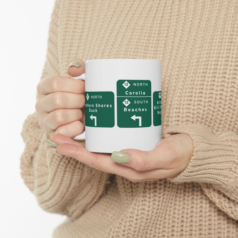 White ceramic coffee mug printed with green Outer Banks highway signs pointing to Corolla, Duck, Kitty Hawk, Kill Devil Hills and Nags Head
